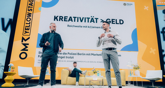 Zwei Polizeibeamte stehen auf einer Bühne des OMR Festivals 2024 und präsentieren ihre Recruiting-Strategie der Polizei Berlin, bei der kreative Kommunikation über teure Kampagnen gestellt wird.