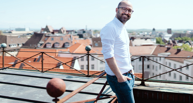 Ein Mann mit Glatze und Brille steht entspannt auf einer Dachterrasse mit Blick über die Dächer einer Stadt, trägt ein weißes Hemd und Jeans, lächelt in die Kamera – Symbolbild für moderne Arbeitsmodelle und New Work.