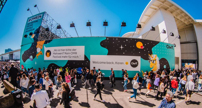 Zahlreiche Besucherinnen und Besucher strömen bei sonnigem Wetter am Eingang des OMR Festivals vorbei. Auf einer riesigen Außenwand steht „#OMR24 – Das Festival für das digitale Universum“, daneben zwei großformatige Aussagen: „Wo ist hier bitte der Mehrwert?“ und „Welcome back, Karen. 🙂“. Die bunte Wandgestaltung zeigt ein Einhorn mit Sonnenbrille und ein stilisiertes Weltall-Motiv. Fotograf: Julian Huke