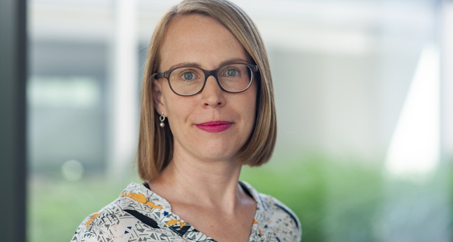 Porträt einer Frau mit kinnlangem blonden Haar, Brille und gemustertem Oberteil. Sie blickt ruhig in die Kamera, im Hintergrund unscharfer Büro- oder Glasfassadenbereich