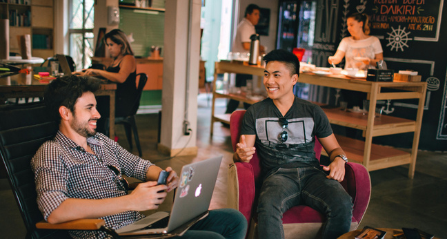 Zwei junge Männer sitzen entspannt in Sesseln in einem modernen Coworking-Space, unterhalten sich lachend und arbeiten an einem Laptop. Im Hintergrund sind weitere Personen an Tischen beschäftigt oder an einer Theke aktiv.