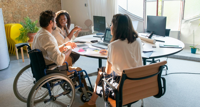 Drei Menschen in einem modernen Büro bei einer Besprechung, eine Person im Rollstuhl. Auf dem Tisch liegen Laptops und Unterlagen.