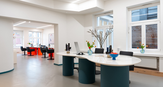 Ein stilvoll eingerichteter, heller Innenraum mit modernen Friseur- und Make-up-Arbeitsplätzen. Im Vordergrund steht ein geschwungener Empfangstisch mit Laptops, Haarprodukten und frischen Blumen in Vasen. Im Hintergrund sind mehrere Stylingplätze an großen Fenstern sichtbar – der Raum wirkt offen, professionell und einladend.