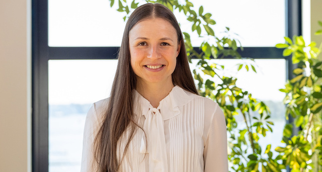 Olga Plotnykova steht lächelnd in einem hellen Büro vor einer Fensterfront mit Pflanzen – Beispiel für erfolgreichen Quereinstieg in die IT durch Netzwerk Chancen.