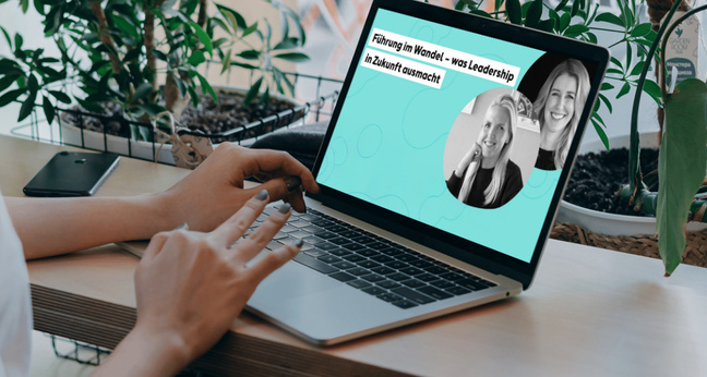 Person sitzt an einem Schreibtisch mit Laptop, auf dem ein Webinar mit zwei Frauen eingeblendet ist. Umgeben von Pflanzen, ruhige Arbeitsatmosphäre.
