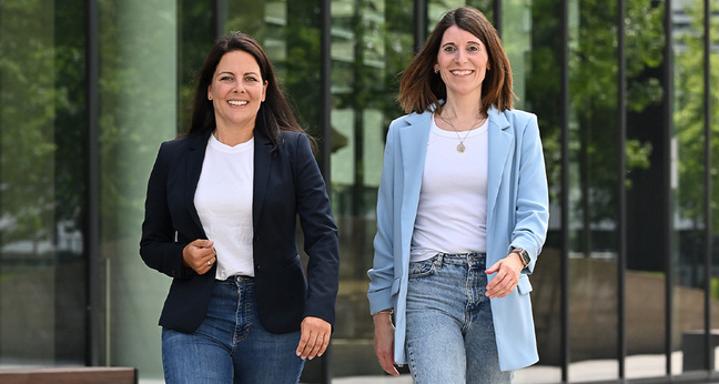 Zwei Frauen gehen lächelnd nebeneinander durch eine moderne Büroumgebung – Gründerinnen der Freelancing-Plattform FreeMOM für berufstätige Mütter.