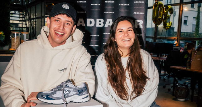Zwei Personen sitzen lächelnd vor einer ADBAKER-Wand, vor ihnen ein einzelner Turnschuh auf einem Podest – Symbol einer unkonventionellen Bewerbung.