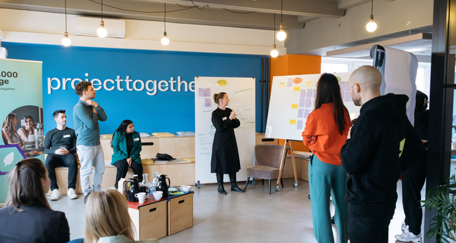 Project Together und AllianzIn einem modernen Workshopraum stehen und sitzen mehrere Personen in angeregtem Austausch vor zwei Whiteboards mit bunten Notizzetteln. Im Hintergrund ist eine große, blaue Wand mit dem Schriftzug „projecttogether“ zu sehen. Links im Bild steht ein Roll-up mit dem Logo der Initiative „10.000 Tage“. Die Szene vermittelt kollaboratives Arbeiten an nachhaltigen Ideen.en: Ein Brainstorming für 10.000 Tage