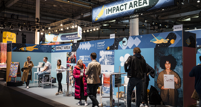 Auf dem OMR Festival 2024 ist ein großzügiger, farbenfroher Bereich mit dem Schild „Impact Area“ zu sehen. Mehrere Stände präsentieren Projekte und Organisationen mit gesellschaftlichem Engagement. Menschen unterhalten sich, informieren sich an den Ständen oder betreuen sie – die Atmosphäre wirkt offen und einladend.