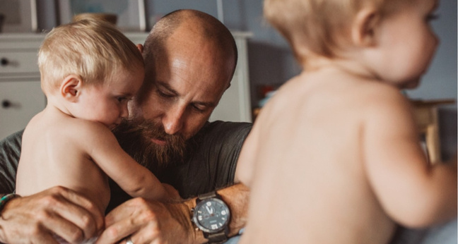 Ein bärtiger Mann hält liebevoll ein kleines Kind auf dem Arm, während ein weiteres Kind im Vordergrund leicht verschwommen zu sehen ist. Die Szene spielt sich in einem gemütlichen, wohnlichen Raum ab und vermittelt Nähe, Fürsorge und familiäre Geborgenheit – Symbol für aktive Elternzeit und moderne Vaterschaft.