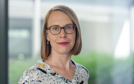 Porträt einer Frau mit kinnlangem blonden Haar, Brille und gemustertem Oberteil. Sie blickt ruhig in die Kamera, im Hintergrund unscharfer Büro- oder Glasfassadenbereich