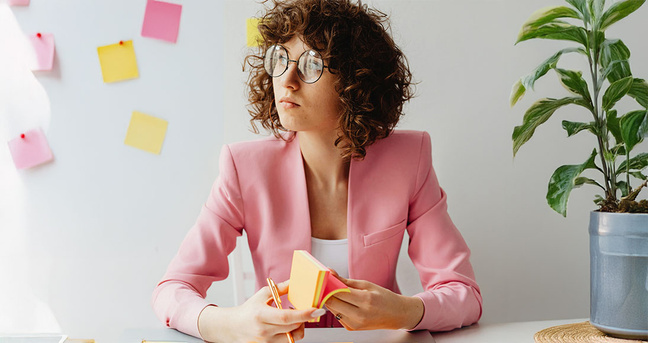 Eine Frau mit lockigem braunem Haar und runder Brille sitzt an einem hellen Schreibtisch. Sie trägt einen rosa Blazer und blickt nachdenklich zur Seite. In ihren Händen hält sie einen Block mit gelben und rosa Klebenotizen und einen Stift. Im Hintergrund ist eine weiße Wand mit mehreren bunten Haftnotizen zu sehen, rechts im Bild steht eine grüne Zimmerpflanze in einem grauen Topf. Die Atmosphäre ist hell, professionell und fokussiert.