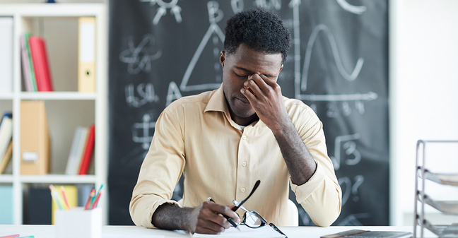 Junge Lehrkraft sitzt erschöpft am Schreibtisch vor einer Tafel mit Formeln – Symbolbild für Überlastung im Referendariat.