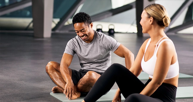 EGYM Wellpass Artikelbild Paid Integration November. Zu sehen sind zwei Menschen in Sportbekleidungen auf einer Matte im Fitnesstudio, die miteinander lachen