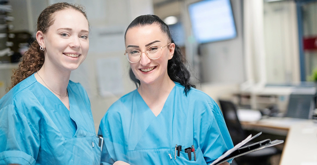 Zu sehen sind zwei Pflegerinnen aus dem Universitätsklinikum Bielefeld. Sie stehen nebeneinander und lächeln in die Kamera. Dabei tragen sie ihre blaue Arbeitskleidung und eine der beiden eine Brille. Im Hintergrund sieht man leicht verschwommen ihren Arbeitsplatz. Sie stehen beispielhaft für die Kolleg*innen, die am Pilot Vier-Tage-Woche in der Pflege teilnehmen.