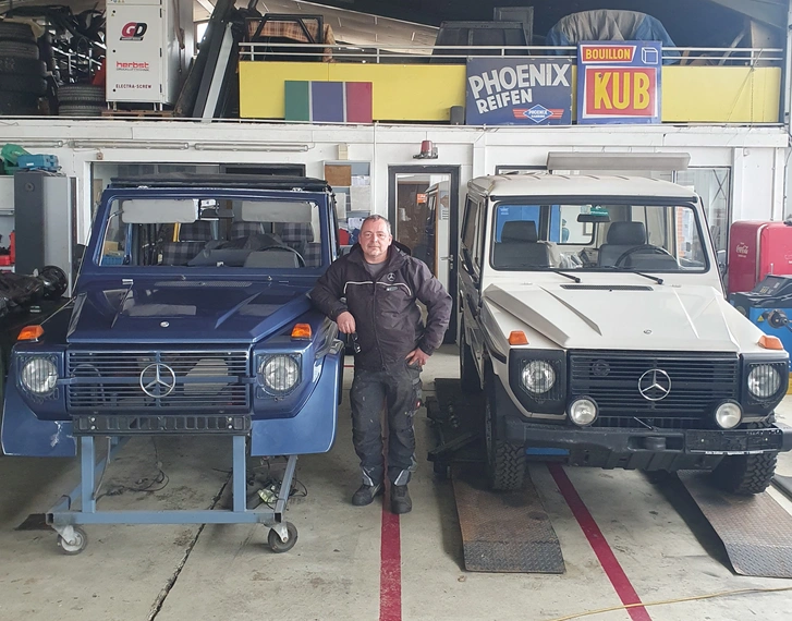In einer Kfz-Werkstatt steht Patrick Lippick in schwarzer Arbeitskleidung zwischen zwei Mercedes G-Klassen. Die Fahrzeuge sind teils restauriert und in Blau und Beige lackiert. Die Aufnahme zeigt eine typische Werkstattumgebung mit Werkzeug, Reifenstapel und Hebebühne – ein authentischer Einblick in den Alltag im Azubi-Recruiting im Handwerk.