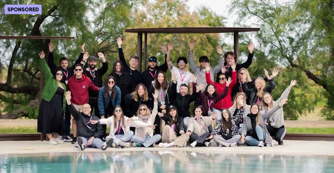 Eine große Gruppe junger Menschen posiert fröhlich vor einer Poollandschaft im Grünen. Einige sitzen, andere stehen hinter ihnen und winken lachend in die Kamera. Die Personen tragen Freizeitkleidung und Namensbänder – die Szene vermittelt Teamspirit, Motivation und eine entspannte Arbeitsatmosphäre.