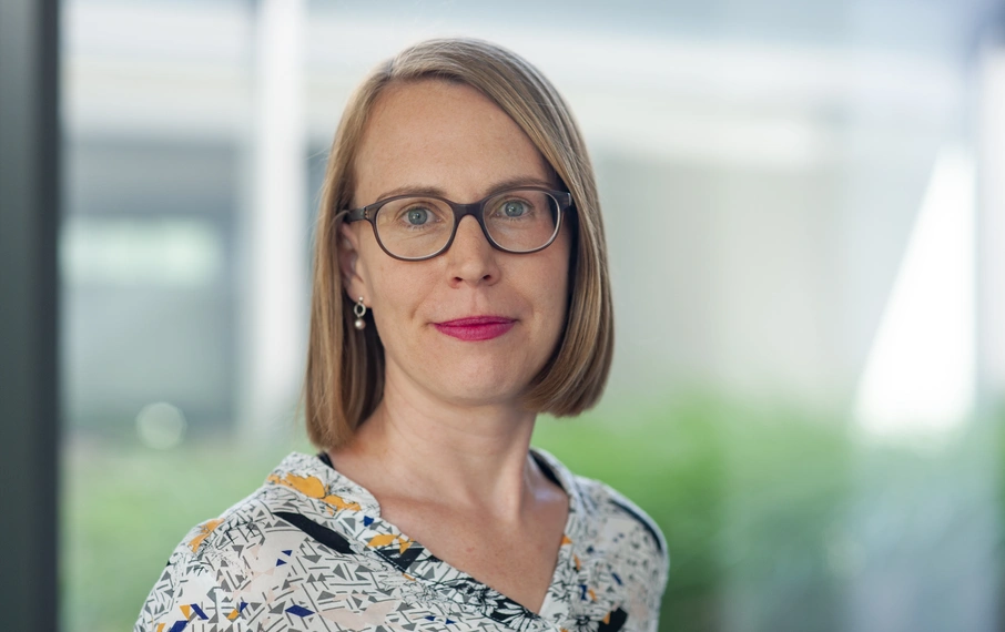 Porträt einer Frau mit kinnlangem blonden Haar, Brille und gemustertem Oberteil. Sie blickt ruhig in die Kamera, im Hintergrund unscharfer Büro- oder Glasfassadenbereich