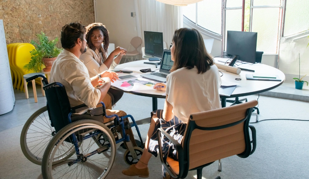 Drei Menschen in einem modernen Büro bei einer Besprechung, eine Person im Rollstuhl. Auf dem Tisch liegen Laptops und Unterlagen.