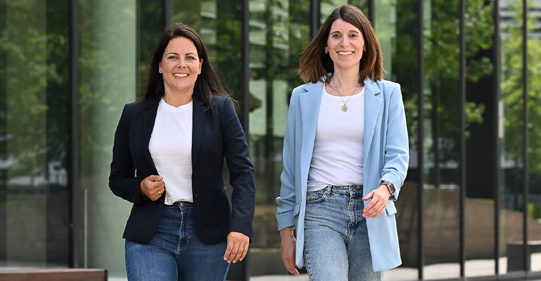 Zwei Frauen gehen lächelnd nebeneinander durch eine moderne Büroumgebung – Gründerinnen der Freelancing-Plattform FreeMOM für berufstätige Mütter.