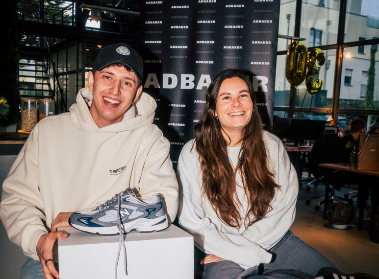Zwei Personen sitzen lächelnd vor einer ADBAKER-Wand, vor ihnen ein einzelner Turnschuh auf einem Podest – Symbol einer unkonventionellen Bewerbung.