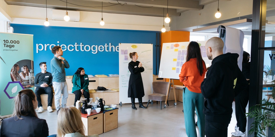 Project Together und AllianzIn einem modernen Workshopraum stehen und sitzen mehrere Personen in angeregtem Austausch vor zwei Whiteboards mit bunten Notizzetteln. Im Hintergrund ist eine große, blaue Wand mit dem Schriftzug „projecttogether“ zu sehen. Links im Bild steht ein Roll-up mit dem Logo der Initiative „10.000 Tage“. Die Szene vermittelt kollaboratives Arbeiten an nachhaltigen Ideen.en: Ein Brainstorming für 10.000 Tage