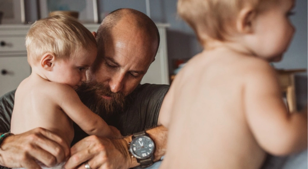 Ein bärtiger Mann hält liebevoll ein kleines Kind auf dem Arm, während ein weiteres Kind im Vordergrund leicht verschwommen zu sehen ist. Die Szene spielt sich in einem gemütlichen, wohnlichen Raum ab und vermittelt Nähe, Fürsorge und familiäre Geborgenheit – Symbol für aktive Elternzeit und moderne Vaterschaft.
