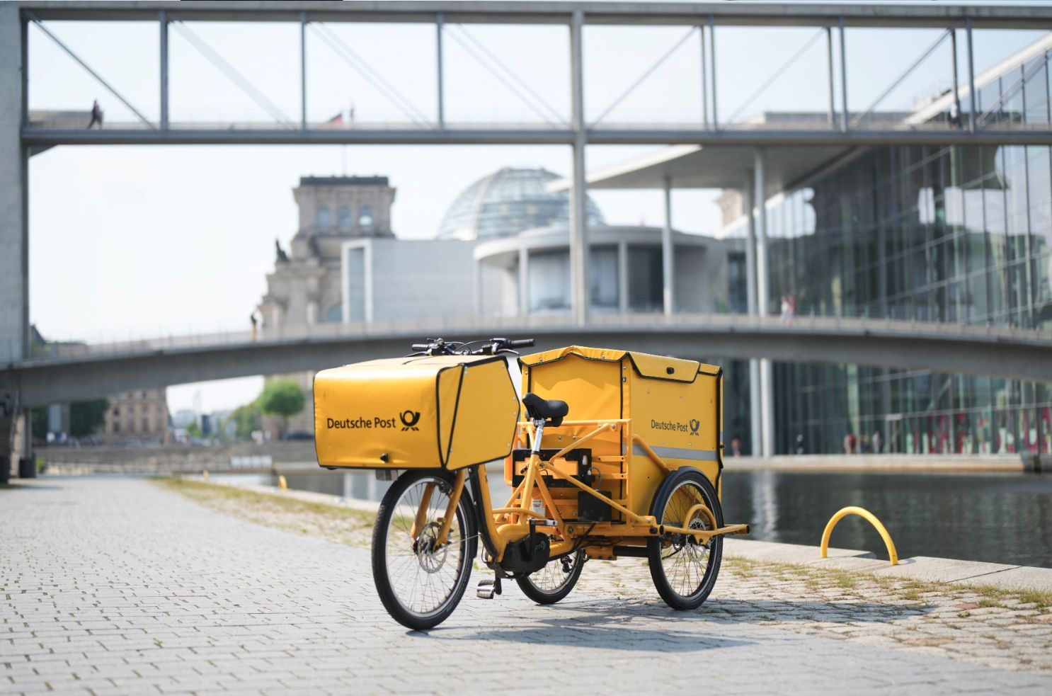 Gelbes E-Trike der Deutschen Post steht am Ufer der Spree in Berlin; im Hintergrund das Regierungsviertel mit Reichstagskuppel und modernen Brücken.