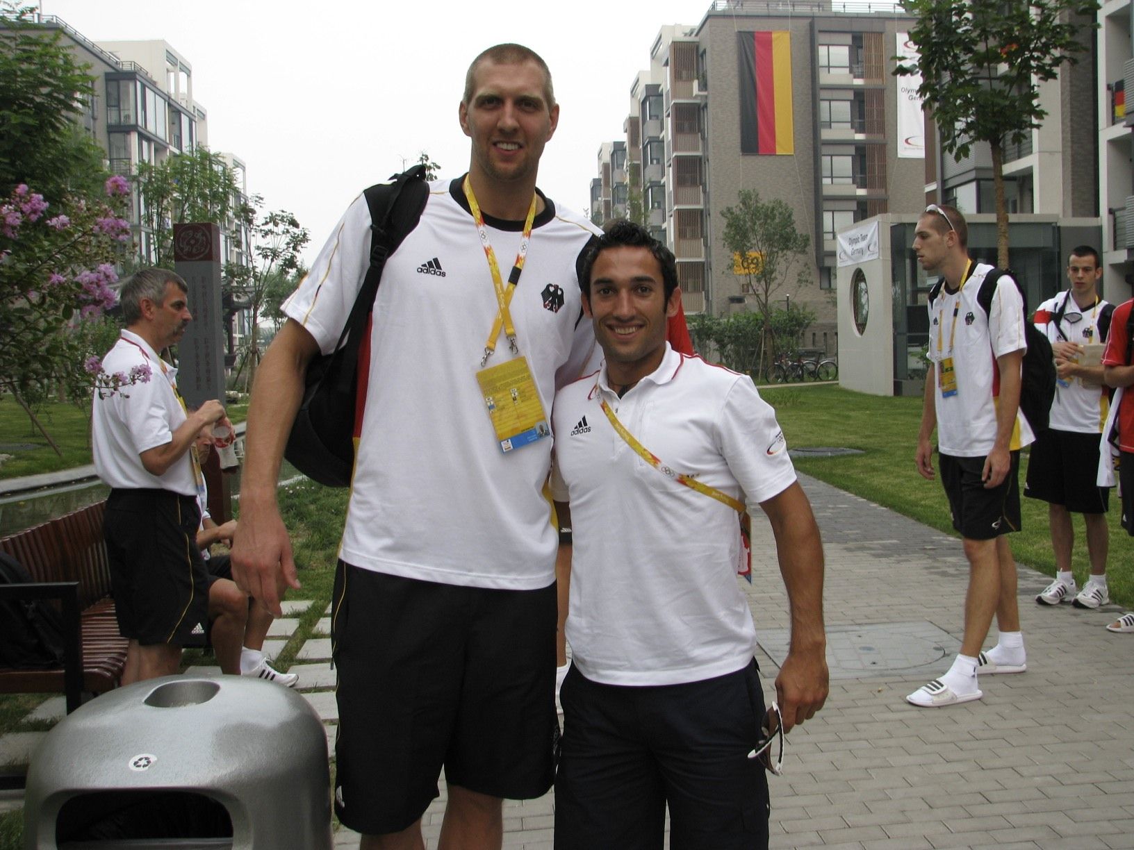 Zwei Männer in Deutschland-Outfits stehen im olympischen Dorf; auffälliger Größenunterschied, im Hintergrund weitere Teammitglieder und deutsche Flaggen.