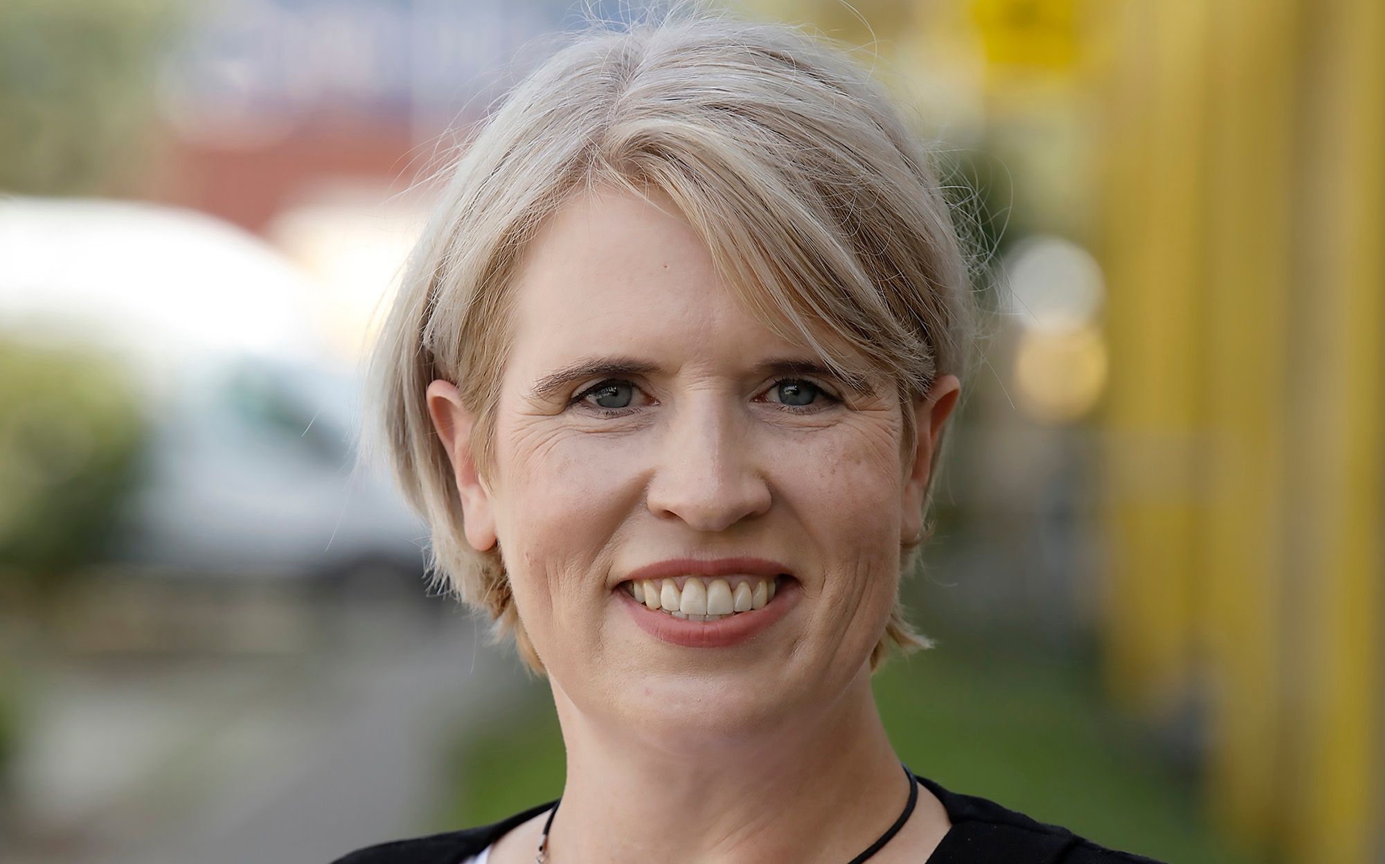 Frau mit kurzem blondem Haar lächelt in die Kamera; unscharfer Hintergrund mit gelben und grünen Farbtönen.