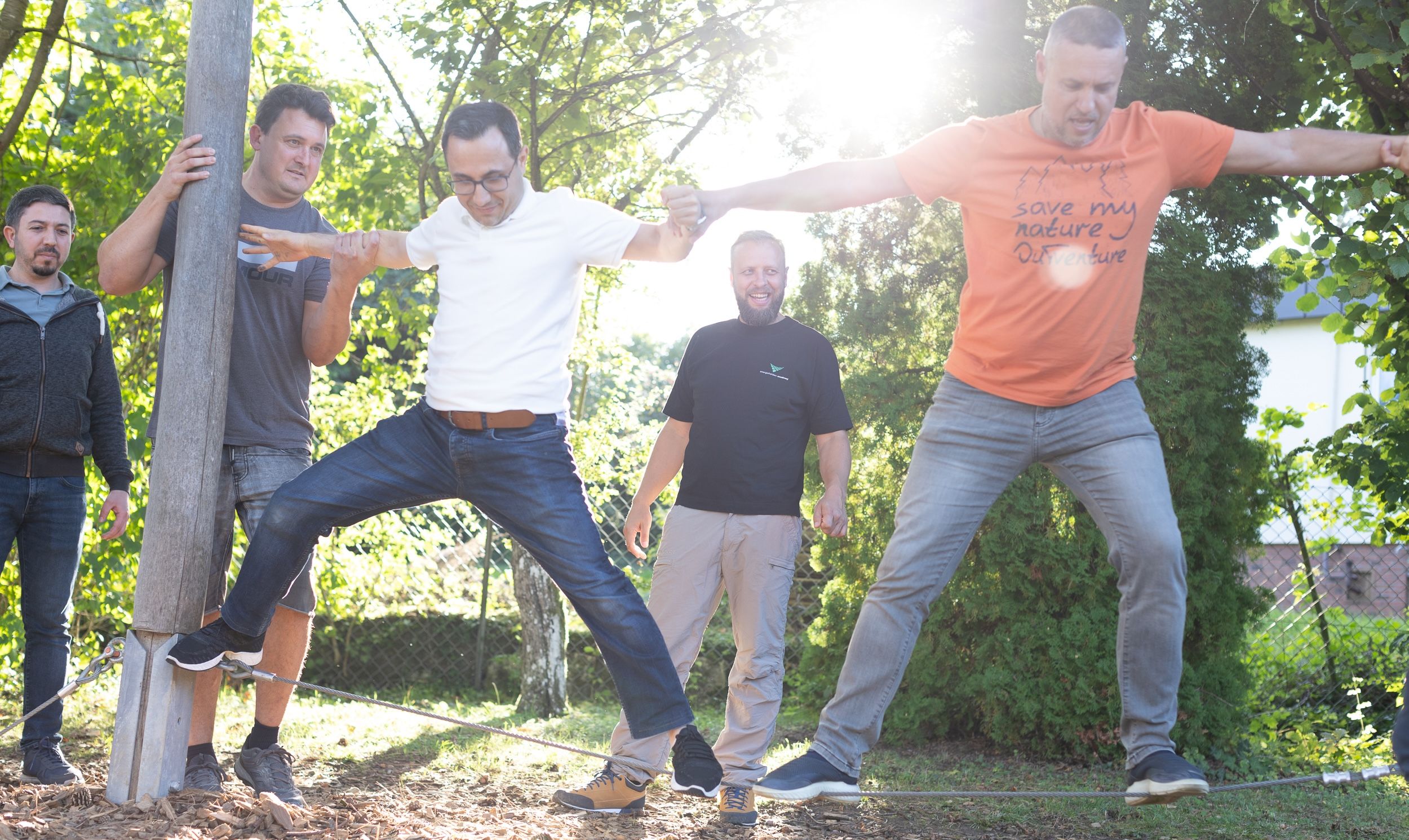 Mehrere Männer balancieren gemeinsam auf einem Seil im Grünen, während sie sich gegenseitig stützen.