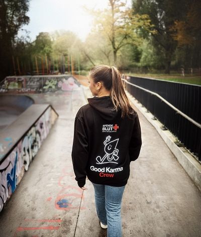 Frau von hinten. Sie trägt einen schwarzen Kapuzenpullover mit der Rückenaufschrift "Good Karma Crew" und dem DRK-Maskottchen, einem Bluttropfen mit Armen und Beinen, der Peace zeigt und lächelt.