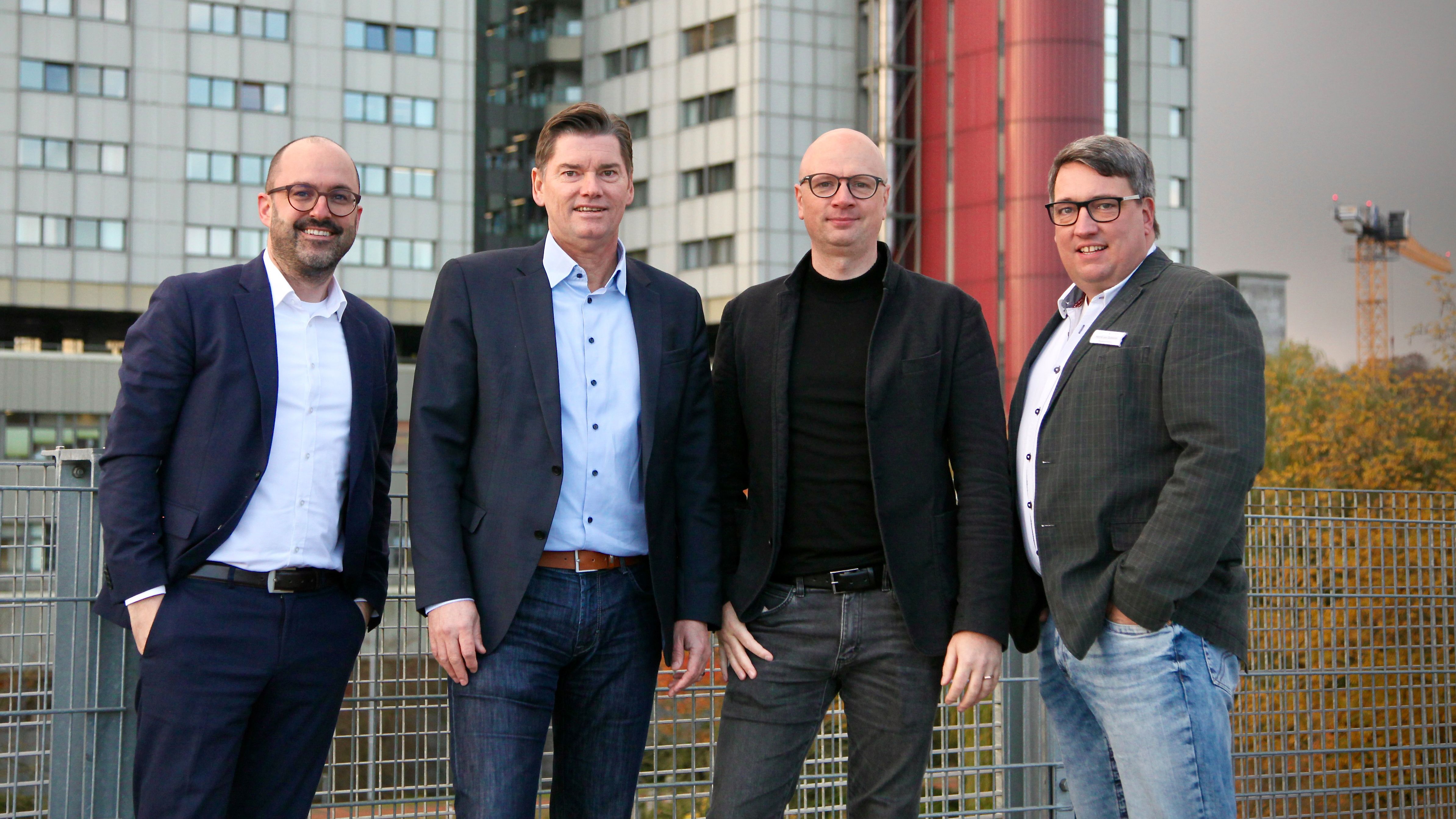 Vier Männer stehen gemeinsam vor einem modernen Klinikgebäude; sie tragen Businesskleidung und lächeln in die Kamera.