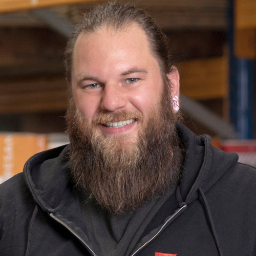 Mann mit Vollbart und langen Haaren, trägt Kapuzenpullover und lächelt in die Kamera; Hintergrund zeigt ein Lager oder Werkstattregal.