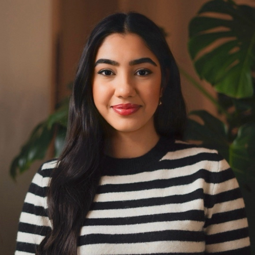 Frau mit langen dunklen Haaren und gestreiftem Pullover lächelt in die Kamera; im Hintergrund eine große grüne Zimmerpflanze.