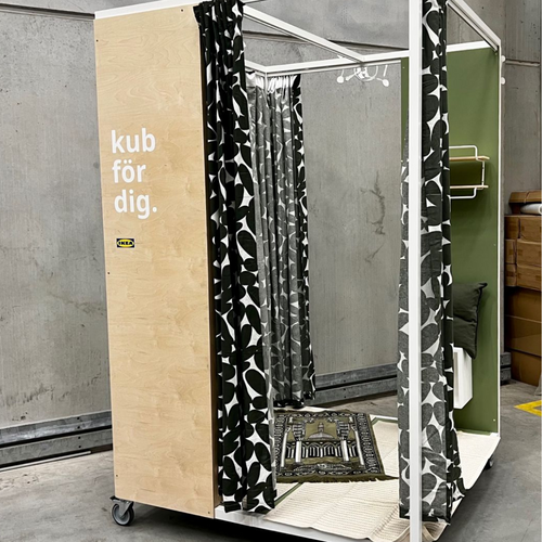 Mobiler, würfelförmiger Rückzugsraum von IKEA mit Vorhängen und Teppich; innen ausgestattet mit Gebetsteppich, Sitzkissen und grüner Wand – konzipiert zur privaten Nutzung, z. B. für Gebete am Arbeitsplatz.