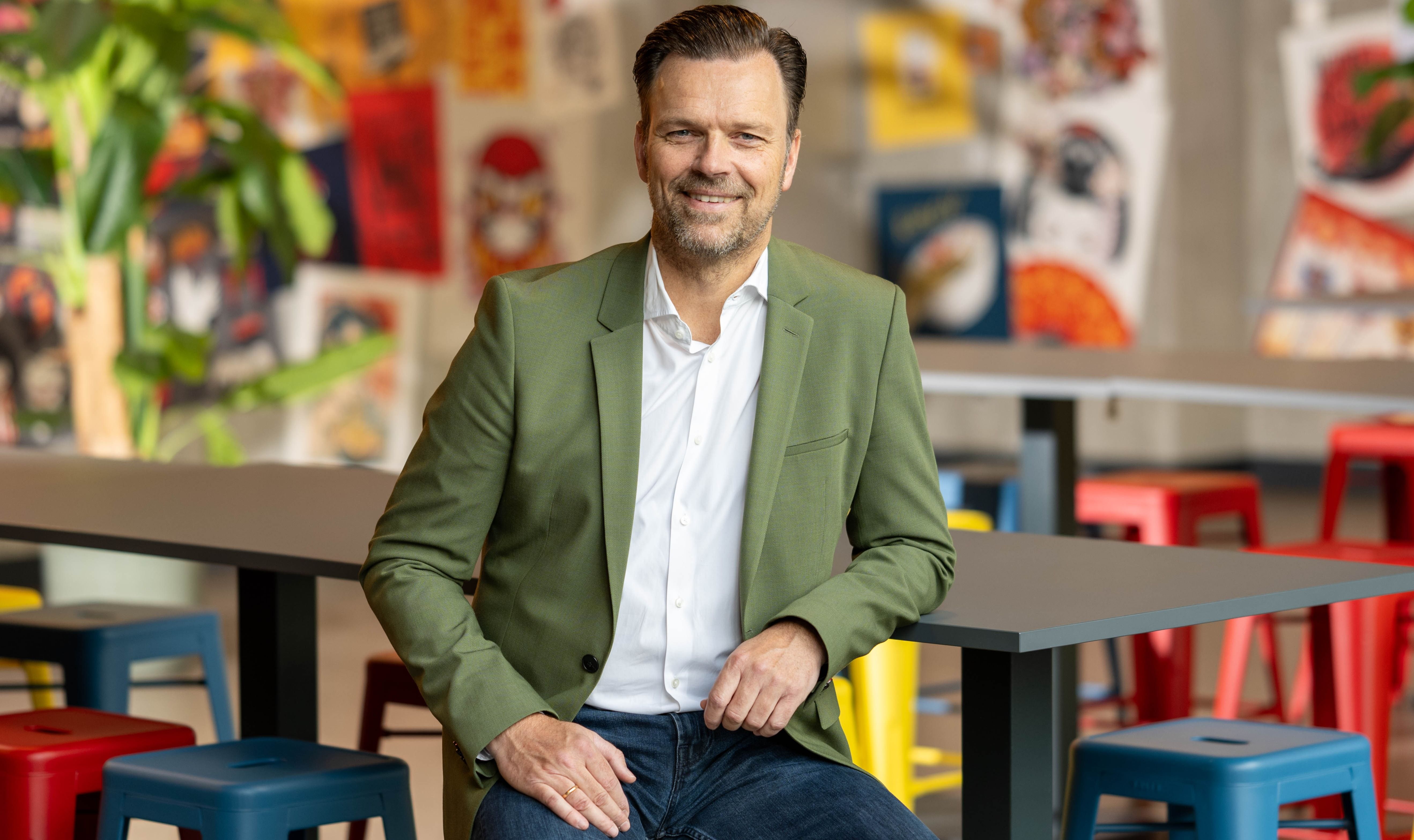 Mann in grünem Sakko sitzt lächelnd an einem Tisch in einem bunt gestalteten Raum mit farbigen Hockern und Wandbildern.