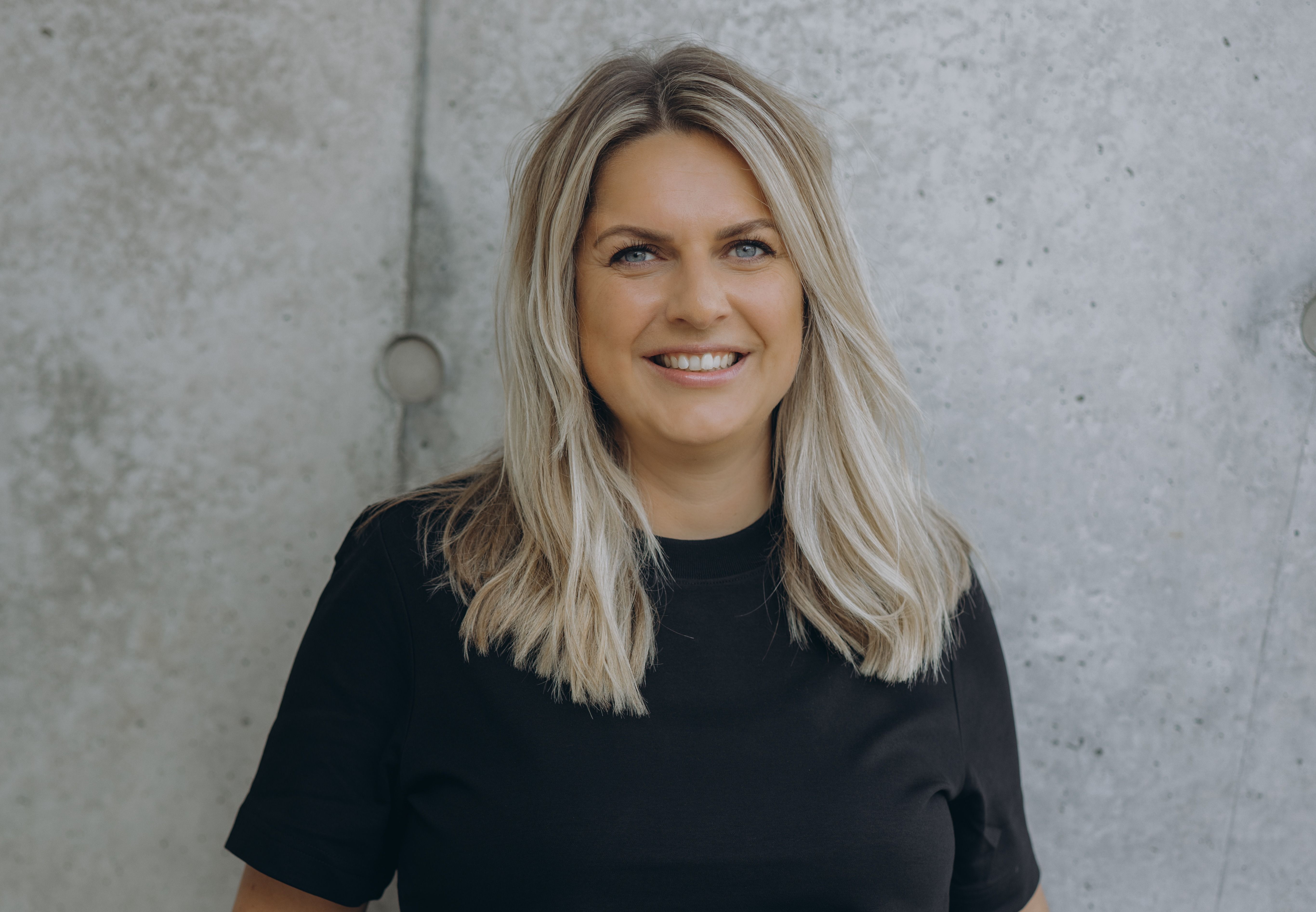 Frau mit langen, blonden Haaren und schwarzem Oberteil lächelt vor einer Betonwand in die Kamera.