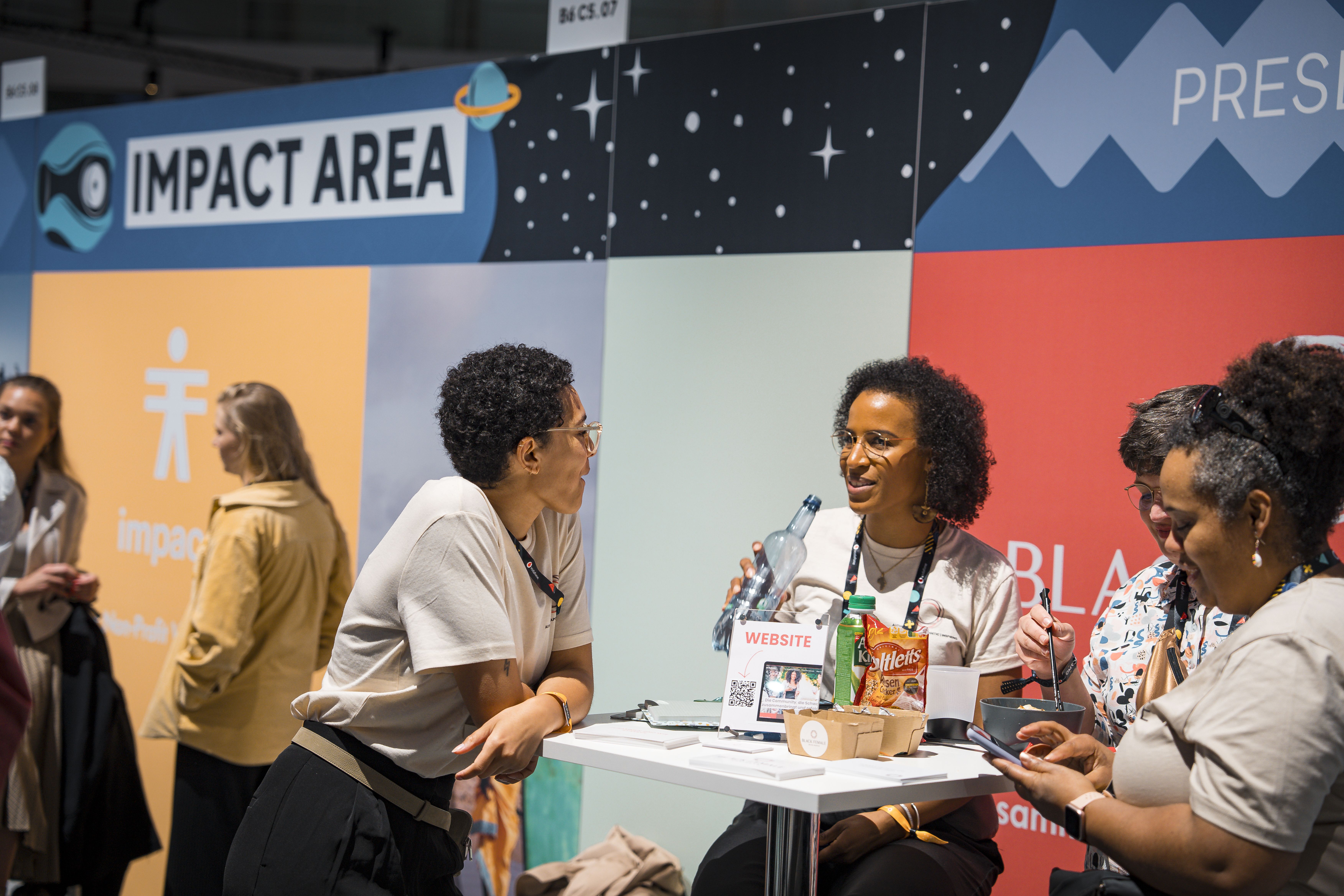 Mehrere Frauen unterhalten sich an einem Messestand in der OMR Impact Area; auf dem Tisch liegen Snacks, Flyer und ein Schild mit der Aufschrift „Website“.
