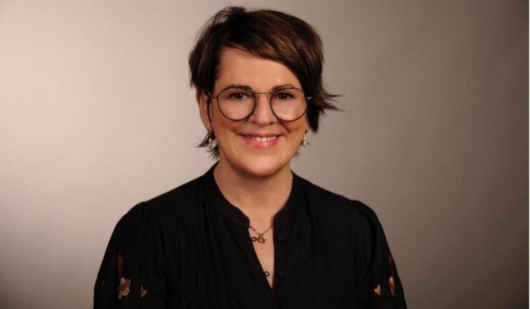 Frau mit Kurzhaarfrisur, Brille und dunkler Bluse lächelt vor neutralem Hintergrund in die Kamera.