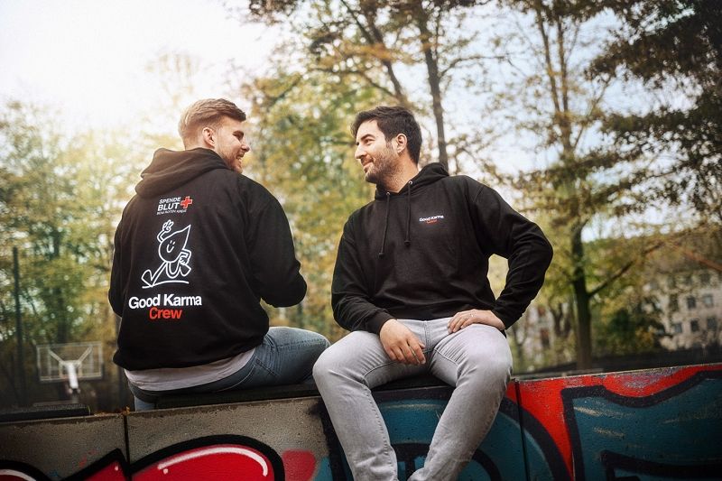 Zwei Männer sitzend, einer von vorn, einer von hinten. Sie tragen schwarze Kapuzenpullover mit der Rückenaufschrift "Good Karma Crew" und dem DRK-Maskottchen, einem Bluttropfen mit Armen und Beinen, der Peace zeigt und lächelt.