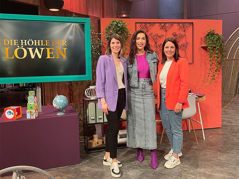 Drei Frauen stehen vor einem „Die Höhle der Löwen“-Schild in einem Studio, bunt gekleidet und lächelnd in die Kamera.