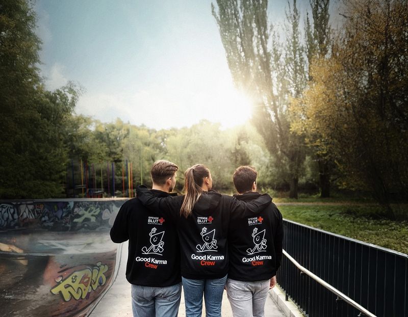 Drei Personen von hinten. Sie tragen schwarze Kapuzenpullover mit der Rückenaufschrift "Good Karma Crew" und dem DRK-Maskottchen, einem Bluttropfen mit Armen und Beinen, der Peace zeigt und lächelt.