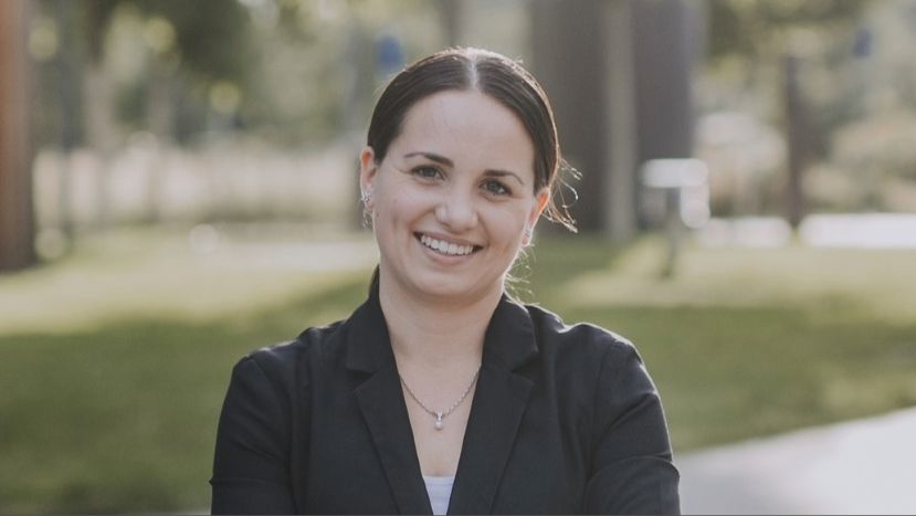 Frau mit dunklen, zurückgebundenen Haaren steht lächelnd im Freien, trägt Blazer und Schmuck; unscharfer Hintergrund mit Bäumen und Weg.